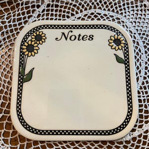 May include: White ceramic notepad with a black and white checkered border. The notepad has a sunflower design on each side and the word "Notes" in black lettering.