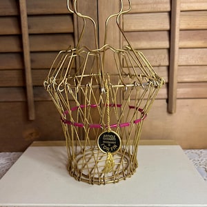 May include: A gold-colored wire basket with a handle and a pink accent ring. The basket has a tag that reads "ARTICLE GARANTI RECOUVERT OR FIN". The basket is sitting on a white surface.