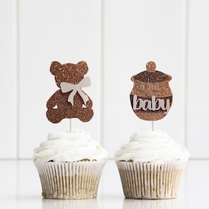 Könnte beinhalten: Zwei Cupcakes mit braunen Glitzer-Toppern. Ein Topper ist ein Bär mit einer weißen Schleife und der andere ist ein Honigtopf mit dem Wort "Baby" darauf.