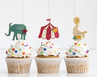 May include: Three cupcakes decorated with white icing and colourful sprinkles. Each cupcake has a different circus-themed topper: an elephant, a tent, and a seal balancing a ball.