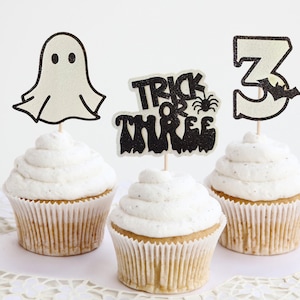 May include: Three cupcakes with white frosting, each topped with a different Halloween-themed decoration. One has a ghost, another says "Trick or Three", and the last has the number 3 with a bat.