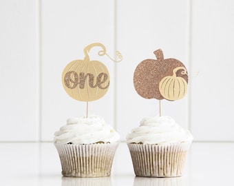 Peut inclure: Deux cupcakes ornés de décorations en forme de citrouille. L'une est dorée avec le mot "one" en paillettes. L'autre est marron avec une plus petite citrouille. Les cupcakes ont un glaçage blanc et sont dans des caissettes en papier brun.