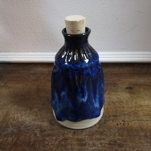 Peut inclure: Une bouteille en céramique faite à la main, avec un glaçage bleu foncé et un bouchon en liège naturel. La bouteille a une forme unique avec une base évasée et un col étroit. Le glaçage bleu a un aspect fluide et texturé.