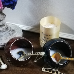 Peut inclure: Deux petits pots à sel en céramique avec des cuillères assorties, l'un brun et l'autre bleu, remplis de sel blanc. Les pots sont sur une surface en bois à côté d'un récipient de couleur crème étiqueté "Petales de Sel à l'Ancienne".