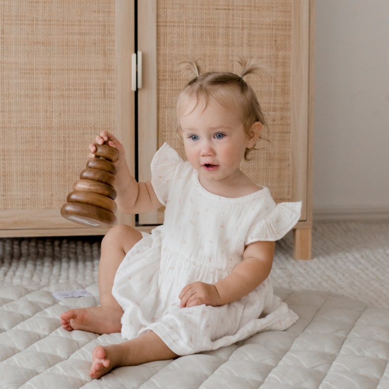 May include: A wooden stacking toy consisting of five smooth, graduated rings on a central post, held by a toddler wearing a white, short-sleeved dress with a subtle pattern. The toy is made of polished brown wood with a natural grain finish.
