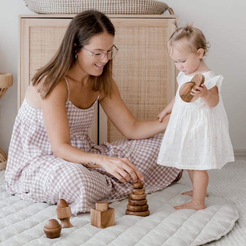 May include: A set of natural wooden stacking toys including a tiered cone, a cube with a cylindrical top, and a small rounded piece with a base. The smooth, polished brown wood pieces are arranged on a quilted surface for tactile play and exploration.