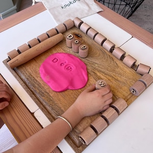 May include: A wooden board with a rolling pin, a set of cylindrical wooden letter stamps, and a pink dough disc imprinted with the letters D, O, and G. A small beige fabric bag with the text kookaroo is positioned at the top of the wooden board.