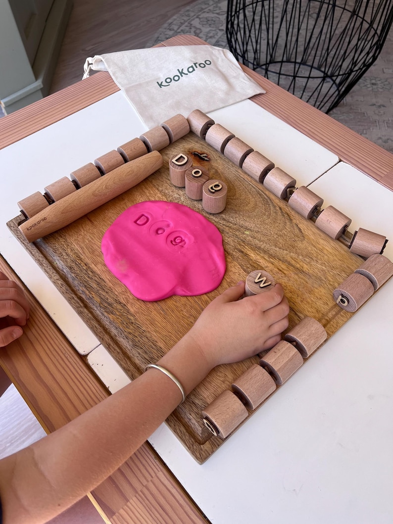 May include: A wooden board with a rolling pin, a set of cylindrical wooden letter stamps, and a pink dough disc imprinted with the letters D, O, and G. A small beige fabric bag with the text kookaroo is positioned at the top of the wooden board.