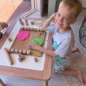 May include: A wooden play tray featuring a rectangular board with two rows of cylindrical wooden letter stamps, a wooden rolling pin, and two flattened pieces of pink and green modeling clay. Small wooden letter stamps are scattered on the table.