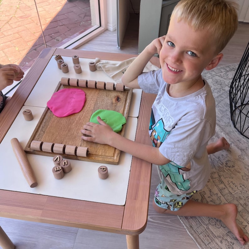 May include: A wooden play tray featuring a rectangular board with two rows of cylindrical wooden letter stamps, a wooden rolling pin, and two flattened pieces of pink and green modeling clay. Small wooden letter stamps are scattered on the table.