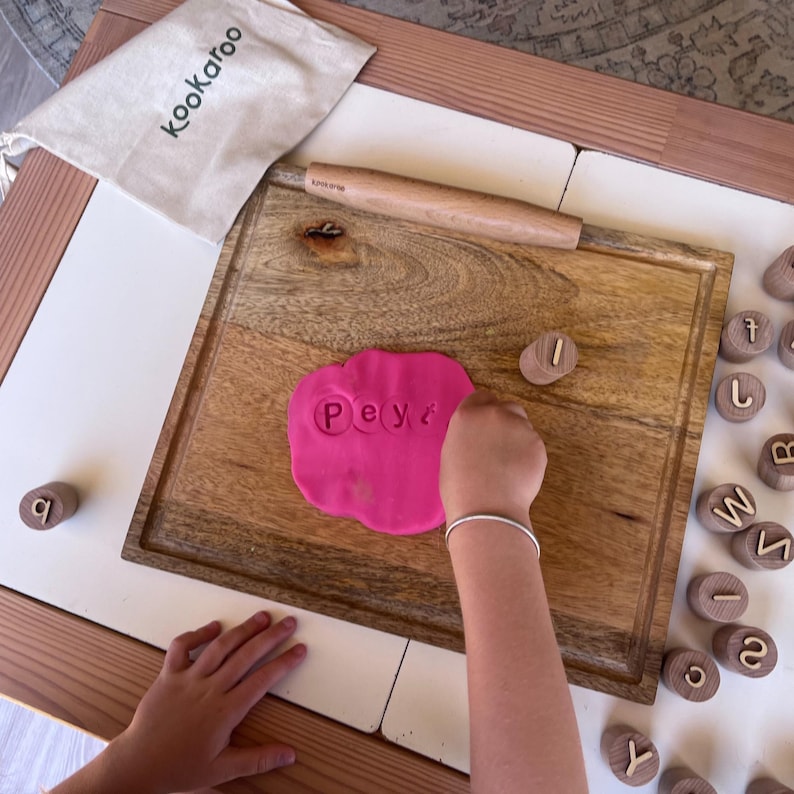 May include: A wooden cutting board with a pink dough slab featuring the imprinted letters P, e, y, and t. A wooden rolling pin rests above the dough. A beige drawstring bag labeled Kookaroo and several small wooden alphabet stamps are nearby.