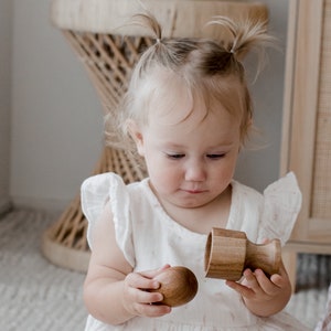 May include: A two-piece wooden toy consisting of a smooth, rounded egg-shaped object and a matching cylindrical cup-shaped base. Both pieces are crafted from natural light brown wood with a visible grain, designed to fit together securely.