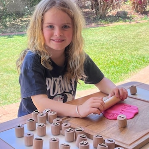 May include: A set of cylindrical wooden stamps featuring embossed letters on the top, arranged on a wooden board. A wooden rolling pin is used to press a letter stamp into a flat piece of pink modeling clay, creating a clear letter impression.