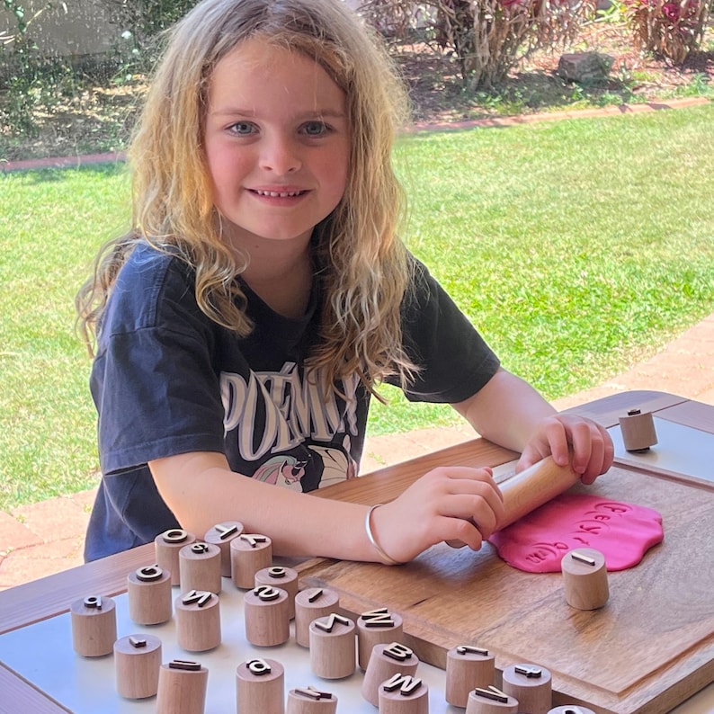 May include: A set of cylindrical wooden stamps featuring embossed letters on the top, arranged on a wooden board. A wooden rolling pin is used to press a letter stamp into a flat piece of pink modeling clay, creating a clear letter impression.