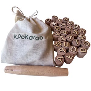 May include: A set of wooden alphabet stamps with a rolling pin and a canvas bag. The bag is beige and has the word "kookaroo" printed on it in green. The stamps are arranged in a pile, and the rolling pin is made of light-colored wood.