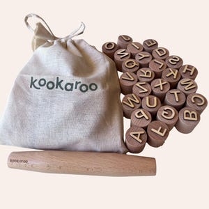May include: A beige drawstring fabric bag with the text kookaroo printed in dark green, a light-colored wooden rolling pin with the text kookaroo, and a collection of cylindrical wooden blocks, each featuring a single letter of the alphabet.