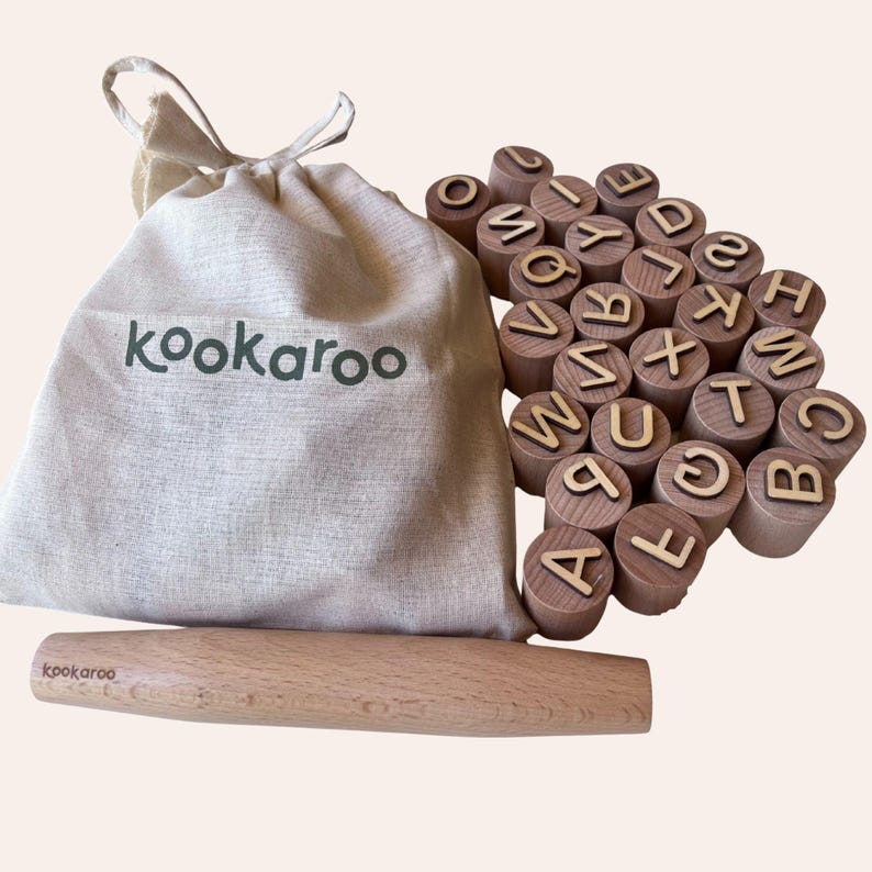 May include: A beige drawstring fabric bag with the text kookaroo printed in dark green, a light-colored wooden rolling pin with the text kookaroo, and a collection of cylindrical wooden blocks, each featuring a single letter of the alphabet.