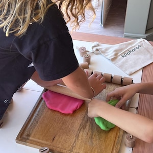 May include: A wooden rolling pin, a rectangular wooden cutting board, and several small wooden cylinders featuring engraved lowercase letters. A beige fabric drawstring bag with the text kookaroo is present, alongside pink and green dough.