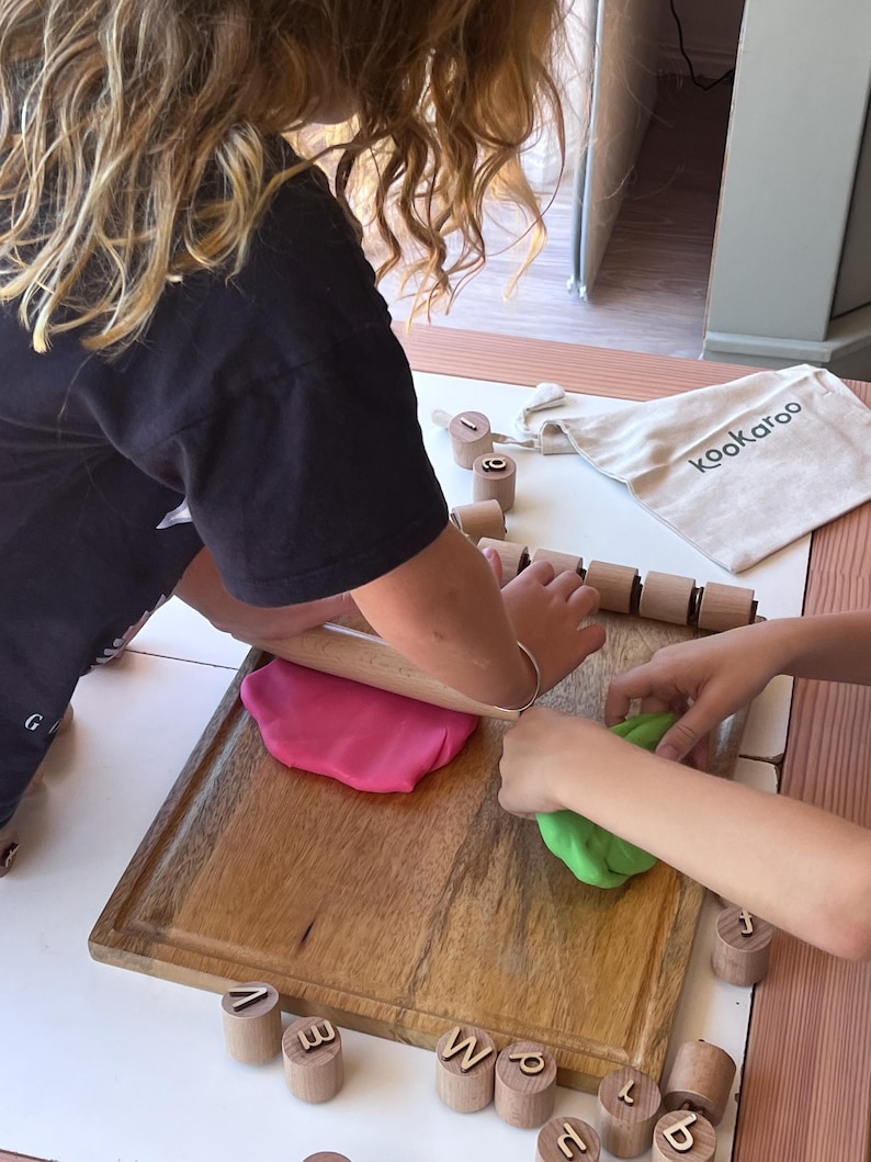 May include: A wooden rolling pin, a rectangular wooden cutting board, and several small wooden cylinders featuring engraved lowercase letters. A beige fabric drawstring bag with the text kookaroo is present, alongside pink and green dough.
