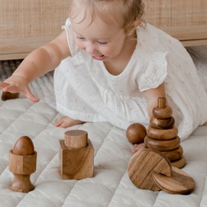 May include: A set of natural wooden toys including a stacking ring tower, a spherical ball, a cube with a cylindrical top, a small egg-shaped piece in a pedestal base, and several flat circular discs with a visible wood grain texture.