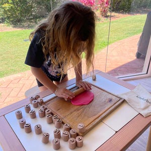 May include: A wooden rolling pin flattening a circular piece of pink dough on a rectangular wooden cutting board, surrounded by a set of cylindrical wooden stamps featuring embossed letters and symbols, with a small beige drawstring bag.