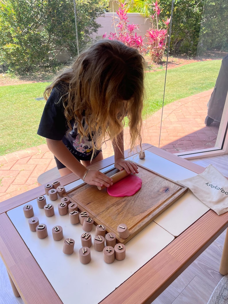 May include: A wooden rolling pin flattening a circular piece of pink dough on a rectangular wooden cutting board, surrounded by a set of cylindrical wooden stamps featuring embossed letters and symbols, with a small beige drawstring bag.