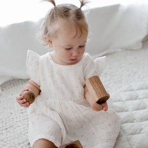 May include: A white dress with flutter sleeves and a subtle pink pattern, paired with a set of natural wooden toys including a small sphere, a cylindrical block, and a square block with a hollow center, all featuring a smooth, polished finish.
