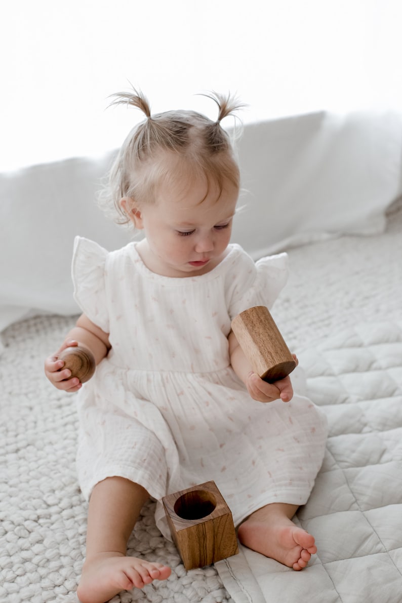 May include: A white dress with flutter sleeves and a subtle pink pattern, paired with a set of natural wooden toys including a small sphere, a cylindrical block, and a square block with a hollow center, all featuring a smooth, polished finish.