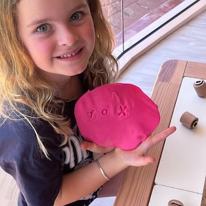 May include: A flat, circular piece of bright pink modeling clay held in a palm. The clay features the lowercase letters f, o, and x pressed into its surface, creating clear indentations. Small wooden cylindrical stamps are visible nearby.