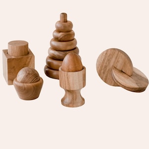 May include: A collection of natural wooden toys including a square block with a cylinder, a stacking ring tower, a smooth egg in a pedestal cup, and a circular puzzle piece. Each item features a polished light brown wood grain finish.