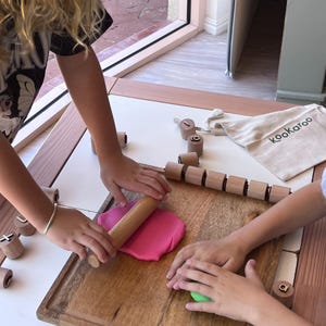 May include: A wooden rolling pin is used to flatten a piece of pink modeling clay on a wooden cutting board. Several small wooden cylinders with engraved letters are scattered around, and a beige fabric bag with the text kookaroo is visible.