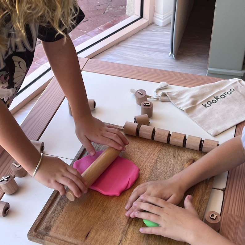 May include: A wooden rolling pin is used to flatten a piece of pink modeling clay on a wooden cutting board. Several small wooden cylinders with engraved letters are scattered around, and a beige fabric bag with the text kookaroo is visible.