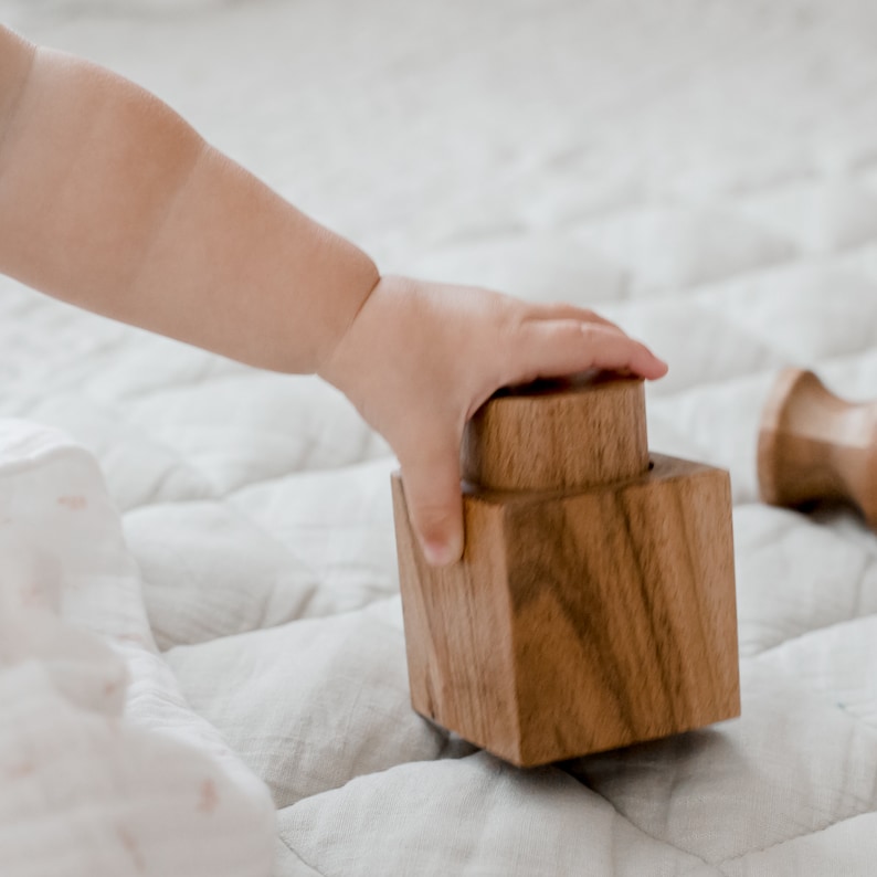 May include: A natural brown wooden block toy featuring a square base and a cylindrical top protrusion. The wood grain is visible across the smooth surface of the object, which is constructed from solid timber with a polished finish and rounded edges.
