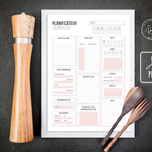 Könnte beinhalten: Eine Holzpfeffermühle mit einem Edelstahlband steht neben einem Tagesplaner mit rosa Akzenten und dem Text "PLANIFICATEUR JOURNALIER". Eine Holzgabel und ein Holzlöffel sind ebenfalls im Bild.
