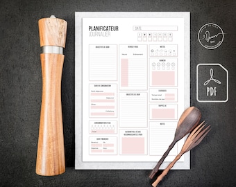 May include: A wooden pepper mill with a stainless steel band stands next to a daily planner with pink accents and the text "PLANIFICATEUR JOURNALIER". A wooden fork and spoon are also in the image.