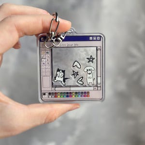 May include: A clear acrylic keychain featuring a retro computer interface design. The keychain includes a black and white cat, a dog, hearts, and stars. The keychain is held by a silver metal clasp.