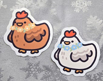 May include: Two cartoon chicken stickers with flower crowns. One chicken is brown with yellow flowers and the other is white with blue flowers.