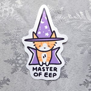Può includere: Un adesivo con un gatto arancione tabby di cartone animato che indossa un cappello da mago viola con stelle e una mezzaluna. Il gatto indossa un mantello viola e il testo "MASTER OF EEP" è sotto il gatto.
