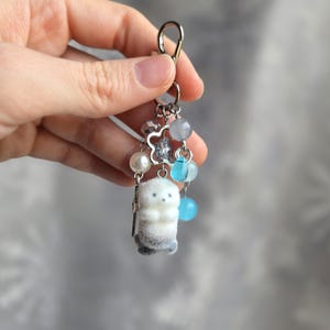 May include: A keychain featuring a small, white plush seal with gray accents. The keychain includes a silver clasp, decorative beads in shades of blue and gray, a pearl, and a silver charm. The seal is the focal point of this charming accessory.