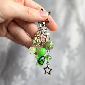 May include: A whimsical keychain featuring a green turtle charm with a black shell design, complemented by green and clear beads, a star-shaped charm, and a star-shaped clasp. The keychain is a charming accessory.