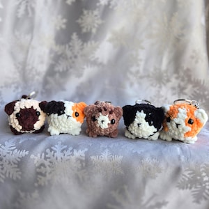 Può includere: Cinque portachiavi a forma di animale all'uncinetto fatti a mano in vari colori, tra cui marrone, nero, bianco e arancione. Ogni portachiavi presenta un piccolo design rotondo a forma di animale con un portachiavi argentato attaccato. I portachiavi sono esposti su uno sfondo morbido a motivi di fiocchi di neve.