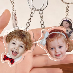 K&ouml;nnte beinhalten: Transparente Acryl-Schl&uuml;sselanh&auml;nger mit individuellen Fotoeins&auml;tzen. Ein Schl&uuml;sselanh&auml;nger zeigt ein l&auml;chelndes Kind mit roter Fliege. Ein anderer zeigt ein kleines M&auml;dchen mit einer blauen Schleife. Die Schl&uuml;sselanh&auml;nger sind an silbernen Ketten befestigt.