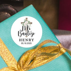May include: A white round sticker with a wooden cross and floral design. The text on the sticker reads "Mi Bautizo HENRY 05.18.2025".