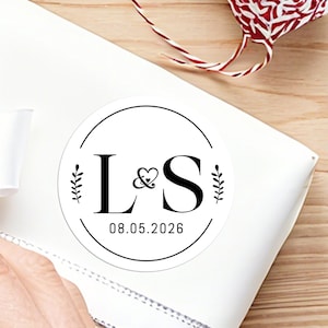 Può includere: Un adesivo rotondo bianco con testo nero e un design minimalista. L'adesivo presenta le iniziali "L & S" con un simbolo a cuore, accenti botanici e la data "08.05.2026" all'interno di una cornice circolare.