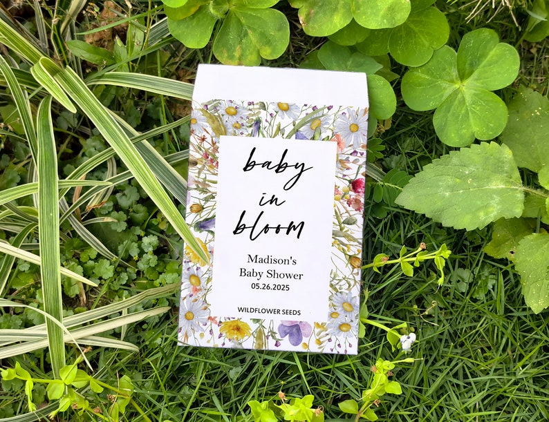 May include: A white seed packet with a floral design and the words "baby in bloom." The packet is surrounded by green grass and foliage. The text "Madison's Baby Shower 05.26.2025 WILDFLOWER SEEDS" is also visible.
