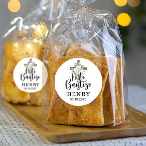 May include: A clear plastic bag filled with small, rectangular pieces of bread. The bag has a white sticker with a cross and flowers, and the text "Mi Bautizo HENRY 05.18.2025".