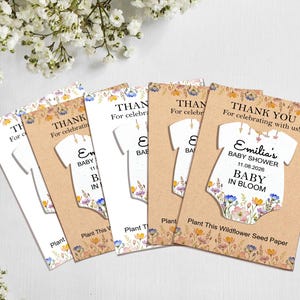 May include: A collection of baby shower thank you cards. Each card features a white onesie shape with floral accents and the text "Emilia's BABY SHOWER 11.08.2026 BABY IN BLOOM". The cards are on a light brown background.