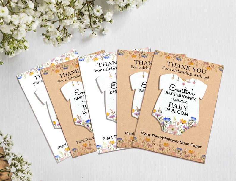 May include: A collection of baby shower thank you cards. Each card features a white onesie shape with floral accents and the text "Emilia's BABY SHOWER 11.08.2026 BABY IN BLOOM". The cards are on a light brown background.