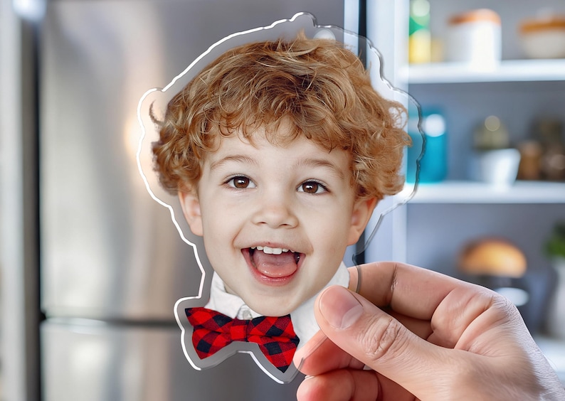 K&ouml;nnte beinhalten: Ein klarer Acrylmagnet mit einem l&auml;chelnden Kind mit lockigem rotem Haar und einer rot-schwarz karierten Fliege. Der Magnet wird vor einem K&uuml;hlschrank aus Edelstahl gehalten.