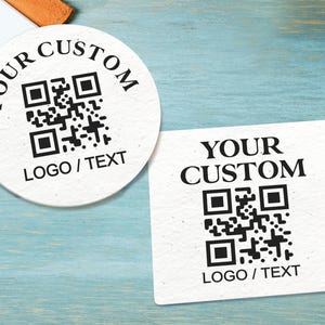 Puede incluir: Dos pegatinas personalizadas, una redonda y otra cuadrada, con un aspecto texturizado blanquecino. Cada pegatina presenta un código QR y el texto "YOUR CUSTOM" y "LOGO / TEXT" en negro, centrado en la pegatina.